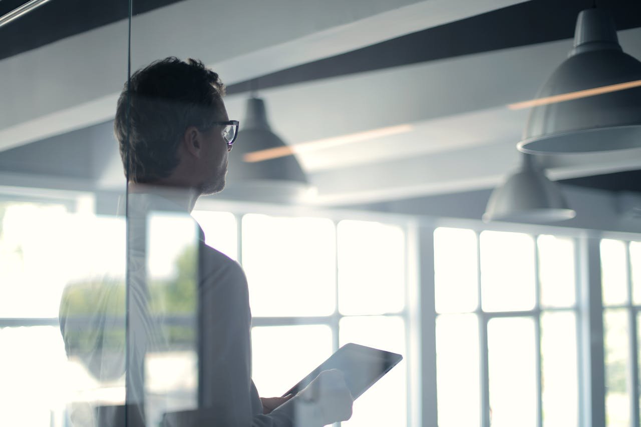 Home A businessman holding a tablet in an office, looking thoughtfully through a glass wall.